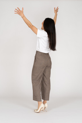 Vista traseira de três quartos de uma jovem feliz de calça e camiseta levantando as mãos