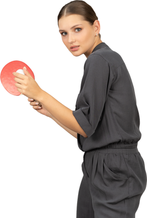 Side view of young woman in a jumpsuit serving tennis ball