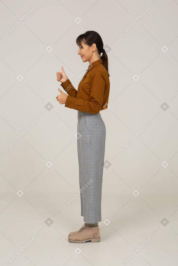 Вид сбоку молодой азиатской женщины в бриджах и блузке показывает палец вверх