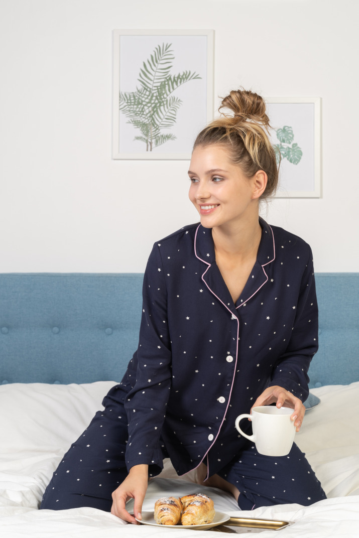 Front view of a young lady in pajamas holding a cup of coffee and some pastries sitting in bed