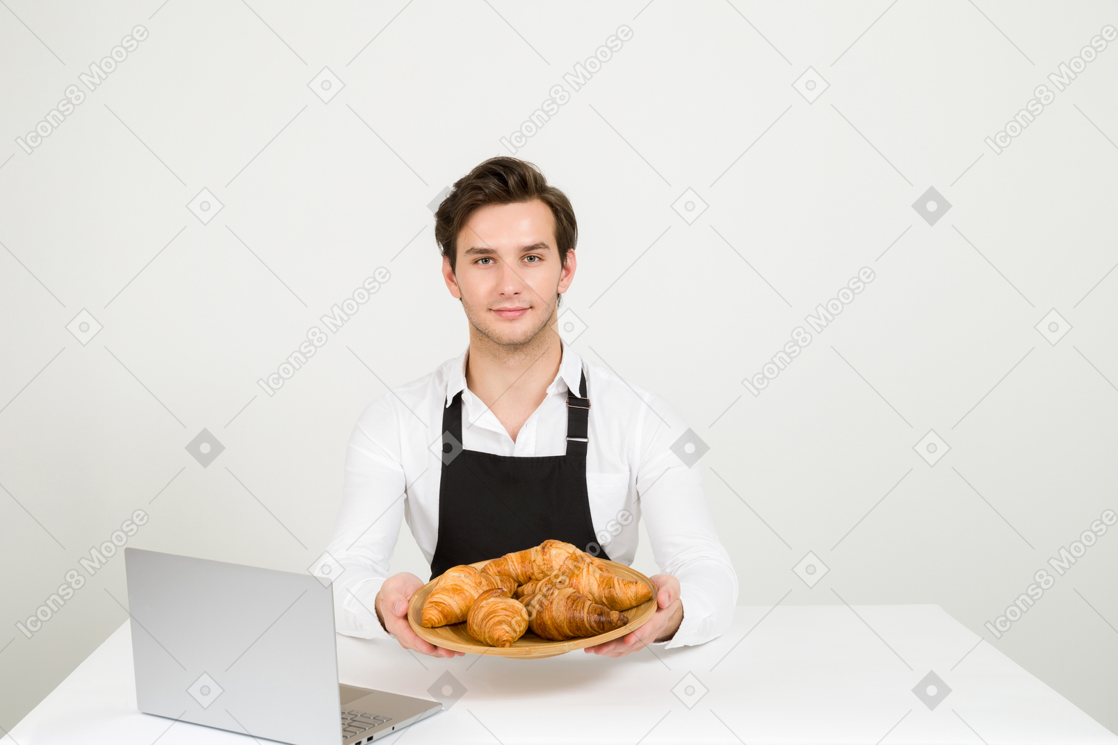 Young baker holding plate of croissants in front of laptop