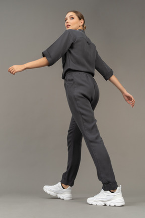 Three-quarter back view of a walking young woman in a jumpsuit looking aside