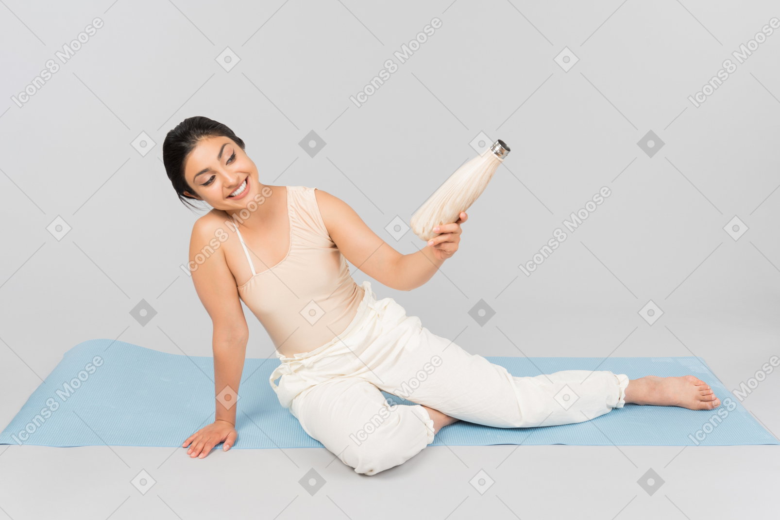 年轻的印度女人坐在瑜伽垫上，拿着运动水壶