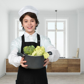 Little boy in a chef's uniform holding a pot with vegetables