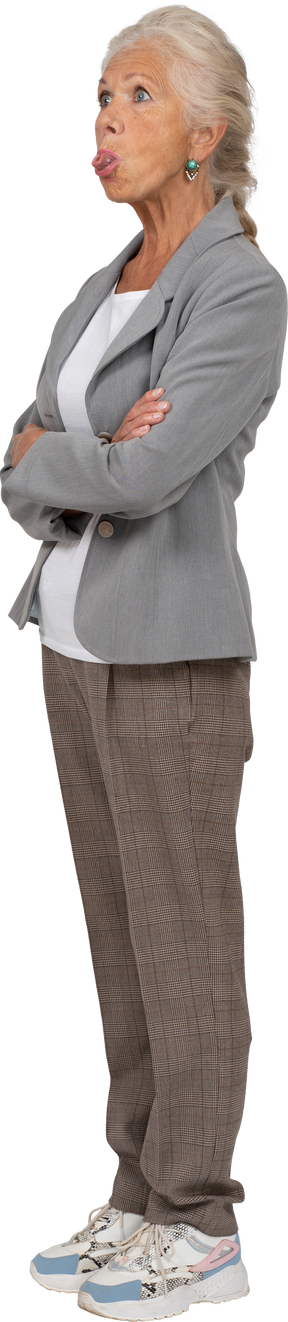 Vista lateral de uma senhora idosa de terno em pé com os braços cruzados e mostrando a língua