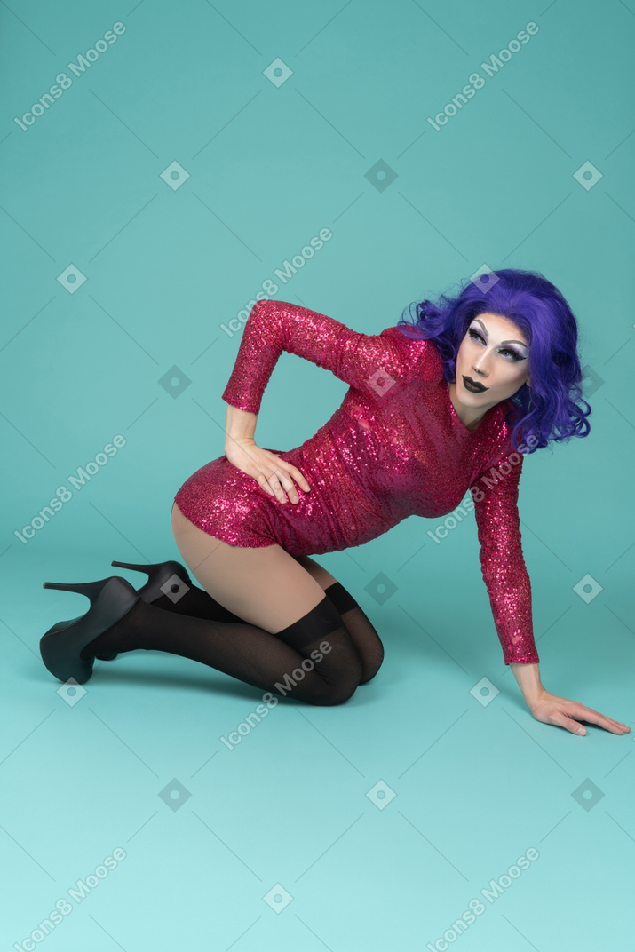 Ganzkörperporträt einer drag queen in rosafarbenem kleid, die mit der hand auf der hüfte auf knien steht