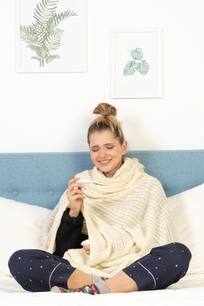 Vue de face d'une jeune femme souriante enveloppée dans une couverture blanche assise dans son lit avec un thermomètre