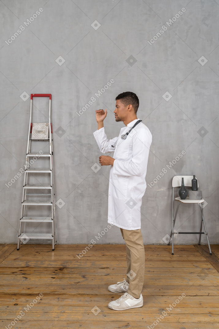 Side view of a young doctor standing in a room with ladder and chair showing a size of something