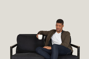 坐在沙发上，拿着一杯咖啡的年轻鬼脸男子的前视图