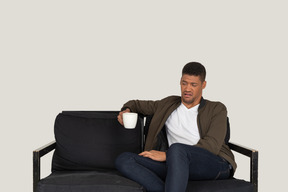 Front view of a young grimacing man sitting on a sofa with a cup of coffee