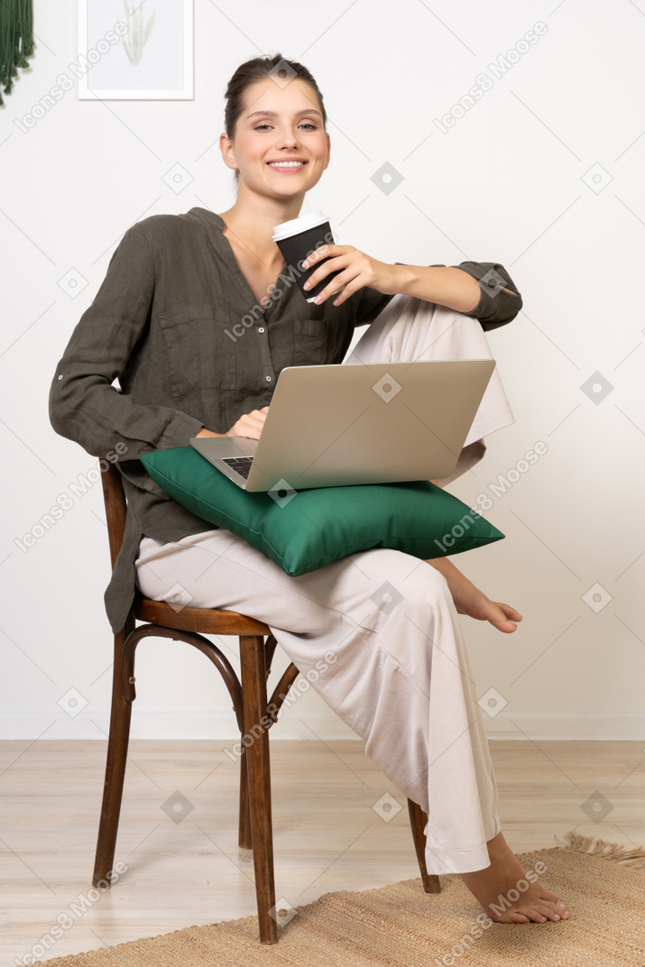 Vista frontal, de, un, sonriente, mujer joven, sentado, en, un, silla, y, tenencia, ella, computadora portátil, y, taza de café