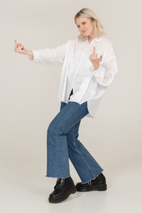 Three-quarter view of a blonde female stepping forward and showing middle finger on both hands and looking aside