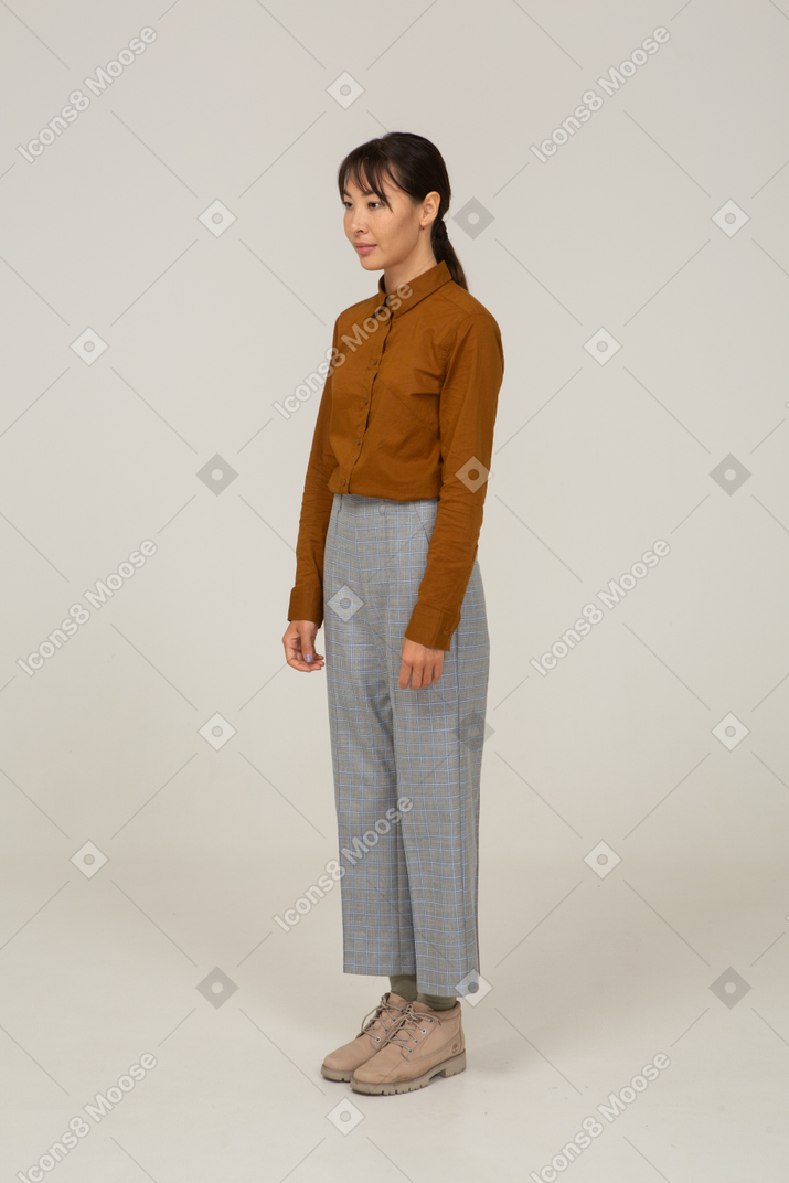 Vista de três quartos de uma jovem mulher asiática de calça e blusa, parada