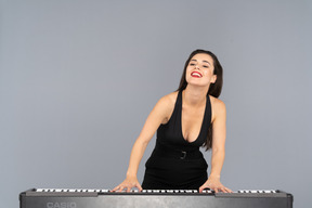 Front view of a pleased young lady in black dress playing the piano while smiling