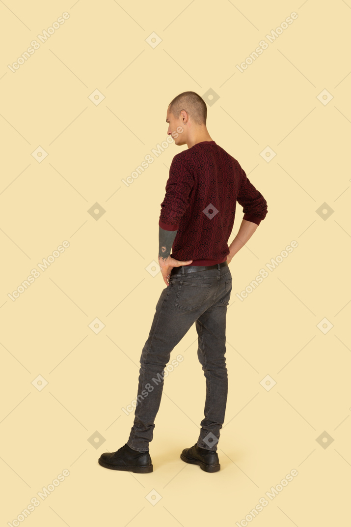 Трехчетвертный вид сзади молодого человека в пуловере, положившего руки на бедра