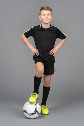 Vista frontal de un niño sonriente en uniforme de fútbol poniendo las manos en las caderas y el pie en la bola