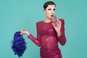 Close-up of a drag queen in pink dress drinking through straw & holding wig