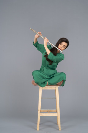 Toute la longueur d'une jeune femme jouant de la clarinette assise, les jambes croisées sur une chaise en bois