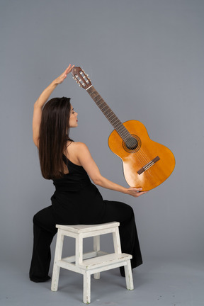 Back view of a young lady in black suit holding the guitar over head and sitting on stool