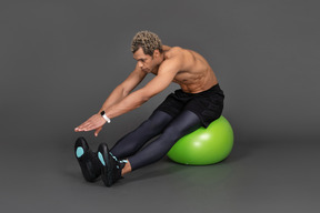 Three-quarter view of a shirtless afro man stretching while sitting on a green gym ball