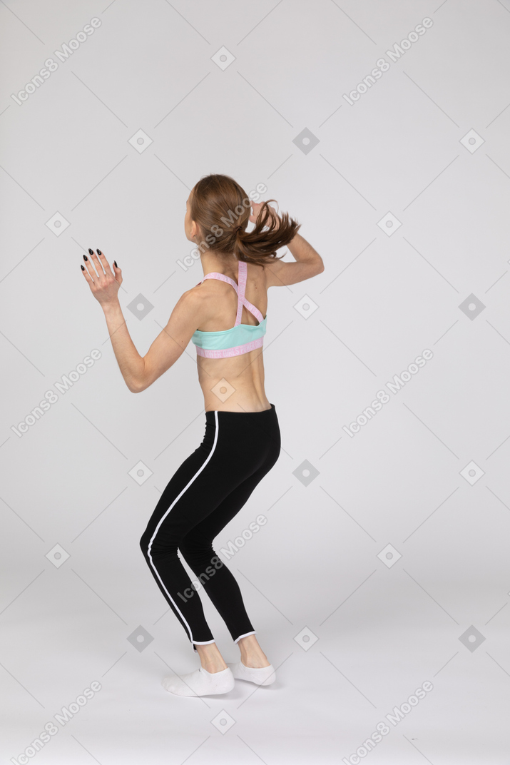 Vista posterior de tres cuartos de una jovencita en ropa deportiva levantando las manos y en cuclillas
