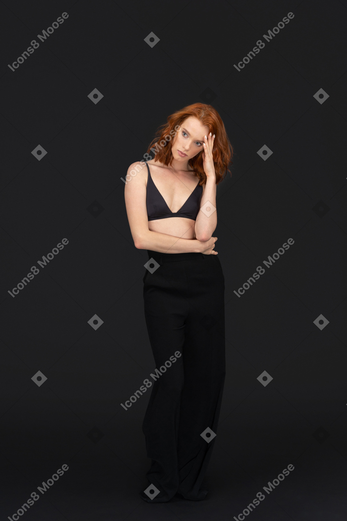 A frontal view of the cute red haired girl posing on the black background and touching her head