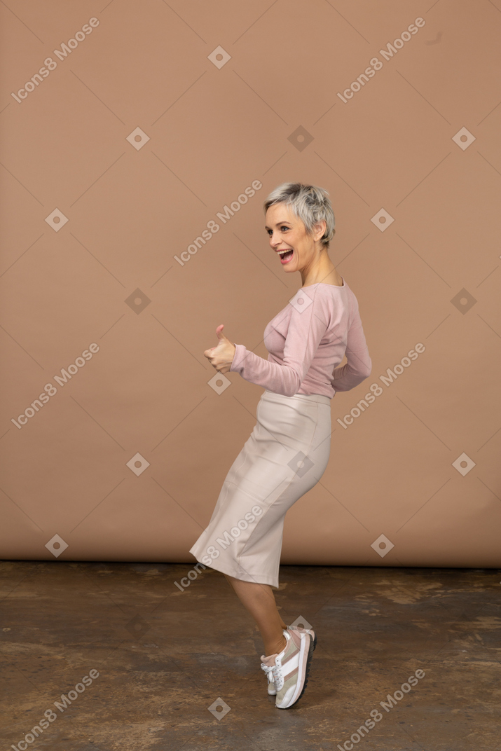 Vista laterale di una giovane donna felice in abiti casual in piedi sulle punte dei piedi e che mostra il pollice in su