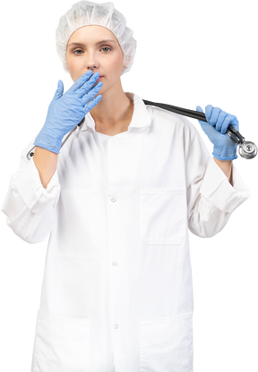 Vista frontal de una joven doctora sosteniendo un estetoscopio y escondiendo la boca