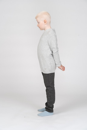 Маленький белокурый мальчик в повседневной одежде стоит и прячет руки за спиной