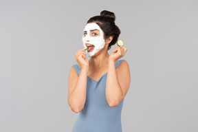 Jeune femme indienne avec masque facial blanc manger des tranches de concombre