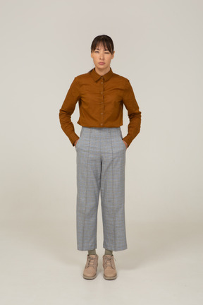 Front view of a young asian female in breeches and blouse narrowing eyes and putting hands in pockets