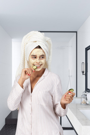 Girl with hair wrapped in towel and mask on her face holding cucumber slices
