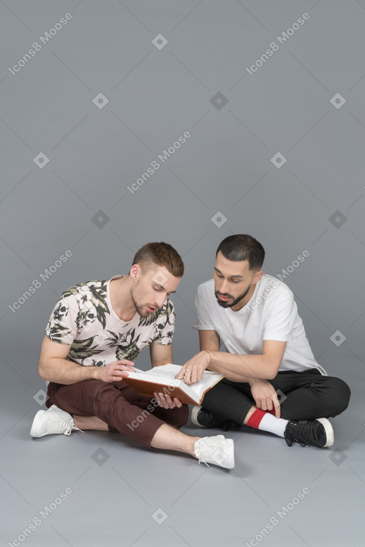 Vue de face de deux jeunes hommes assis sur le sol et étudiant un livre