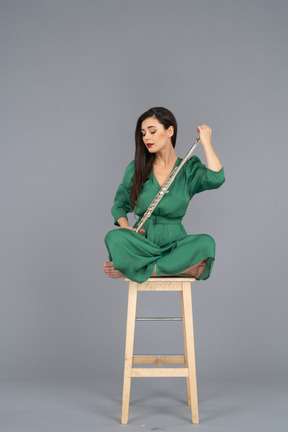 Corpo inteiro de uma jovem olhando para o clarinete sentada com as pernas cruzadas em uma cadeira de madeira