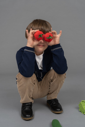 Little boy holding two tomatoes in front of his eyes while squatting