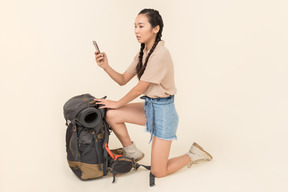 Outraged young asian woman standing near backpack and taking picture with smartphone