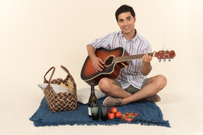 Young caucasian guy sitting near picnic basket on the blanket and playing guitar