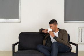 Vista de três quartos de um jovem sentado em um sofá enquanto bebe café