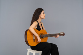 Vue de trois quarts d'une jeune femme assise en costume noir à jouer de la guitare