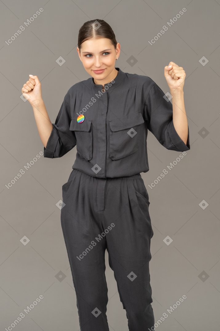 Front view of a young woman in a jumpsuit with lgbt pin raising hands & clenching fists