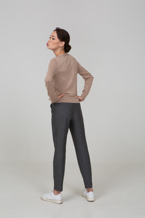 Back view of a young lady in pullover and pants sending an air kiss and putting hands on hips
