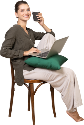 Three-quarter view of a smiling young woman sitting on a chair and holding her laptop & coffee cup