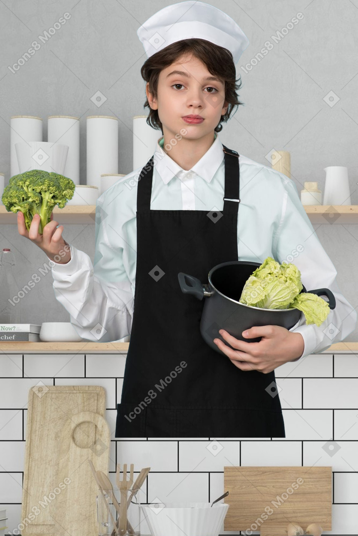 A boy in a chef's outfit holding broccoli