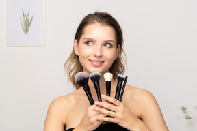 Vista frontal de una mujer joven sonriente sosteniendo pinceles de maquillaje y cabeza inclinada