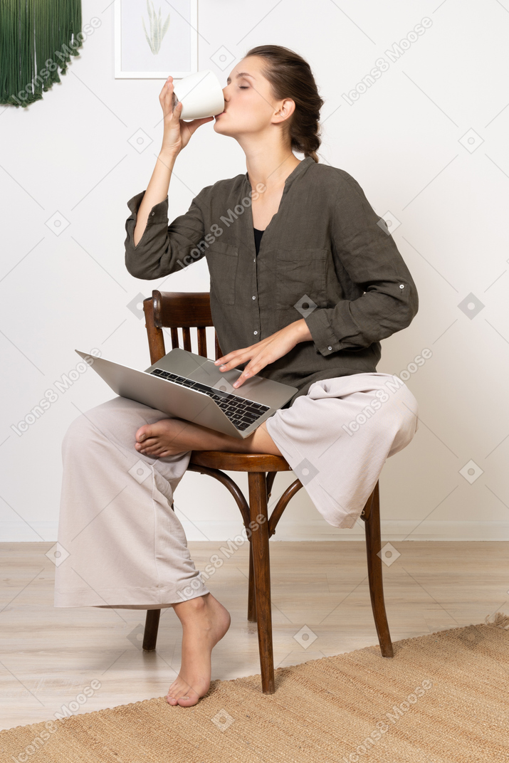 Front view of a young woman wearing home clothes sitting on a chair with a laptop & drinking coffee