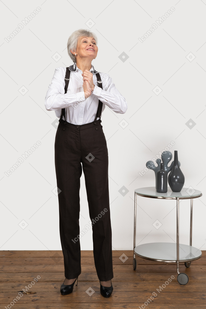 Front view of an old pleased lady in office clothing holding hands together while looking up