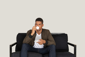 Front view of a young dreaming man sitting on a sofa while drinking coffee