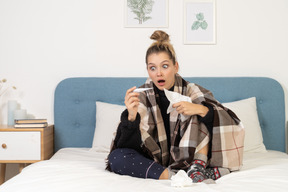 Vista frontal de uma jovem doente em choque, de pijama, enrolada em um cobertor xadrez, verificando sua temperatura