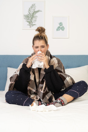 Front view of an ill young lady in pajamas wrapped in checked blanket in bed blowing her nose