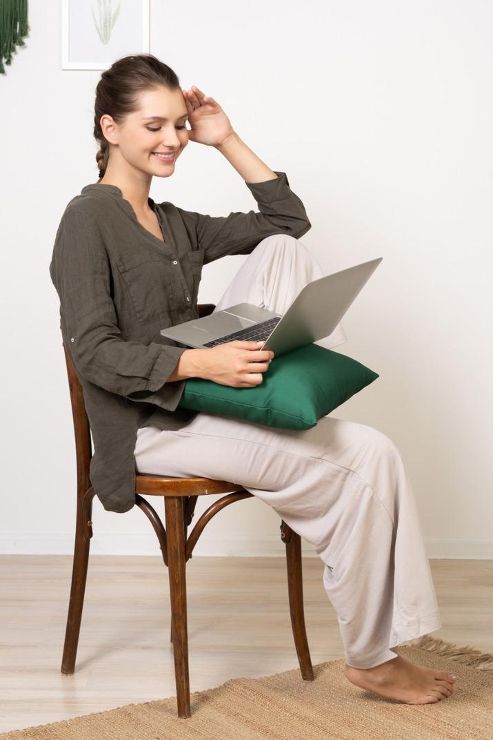 Side view of a young woman wearing home clothes sitting on a chair with a laptop